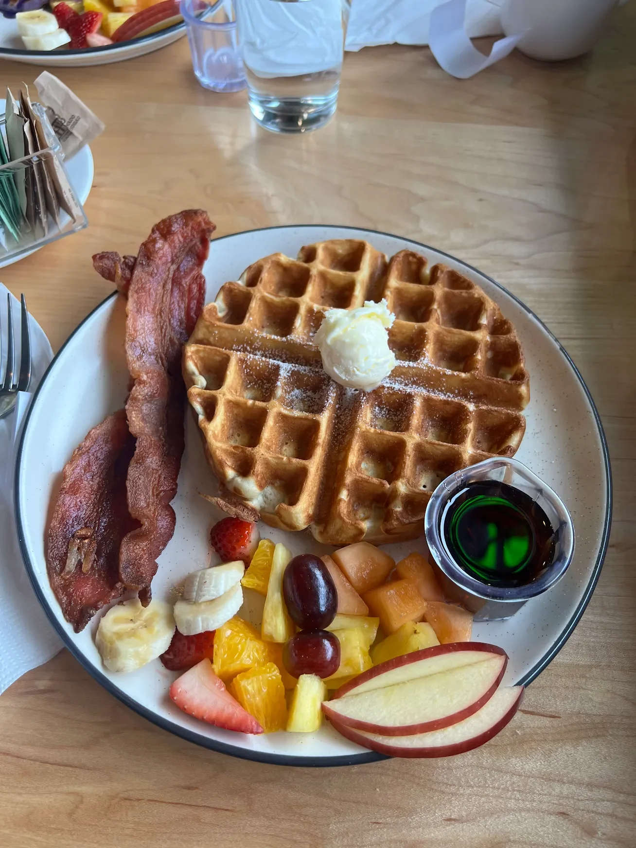 Golden waffle with butter, crispy bacon, fresh fruit, and maple syrup served on a breakfast platter
